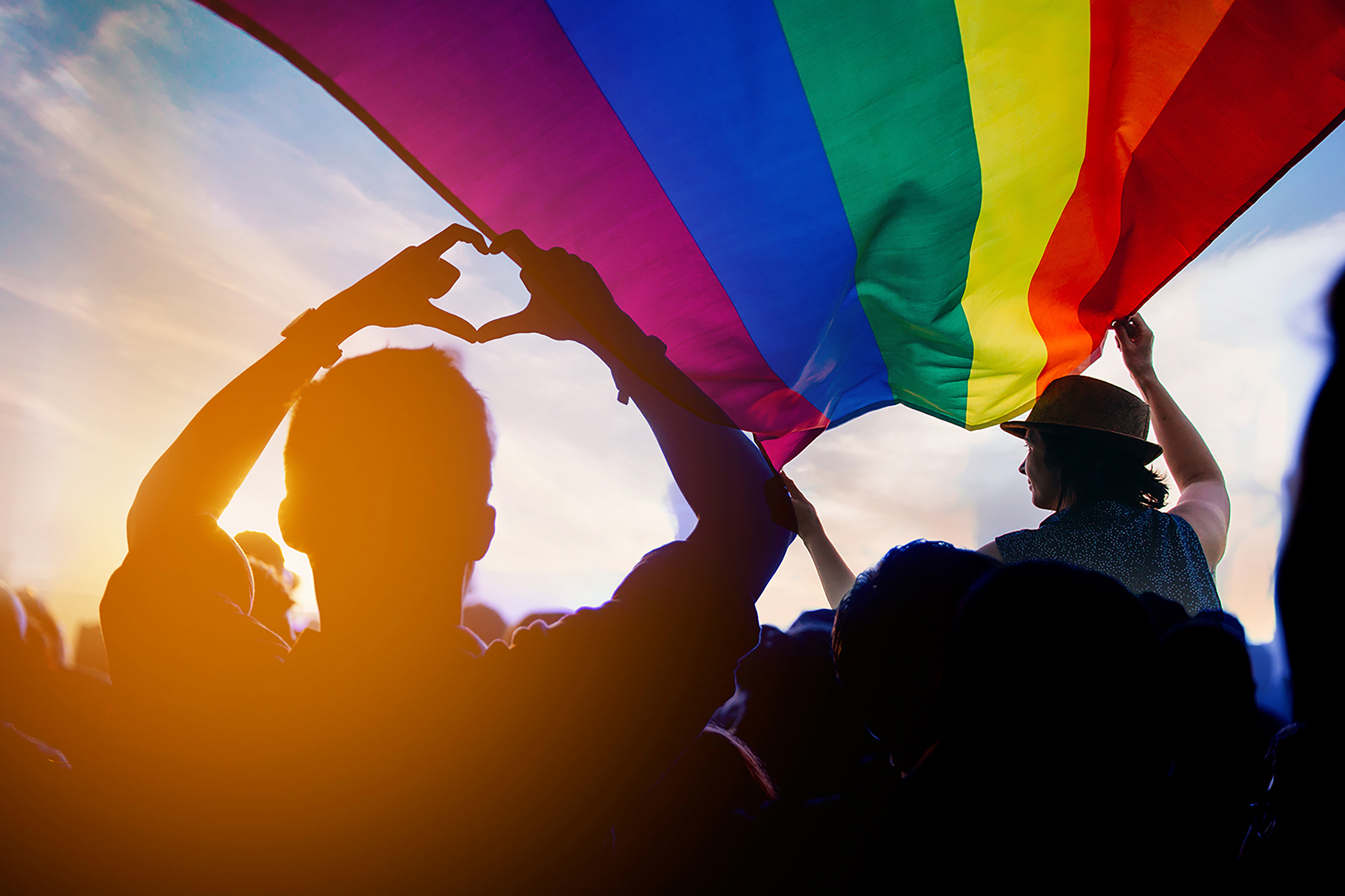 Young people in silhouette carry a rainbow flag under a blue ski.