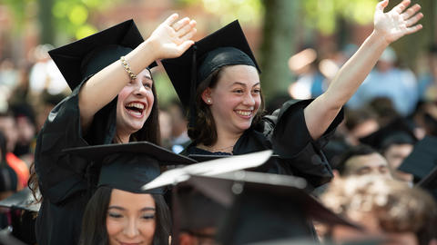 Two graduates in caps and gowns wave at family members before Baccalaureate Services 20026