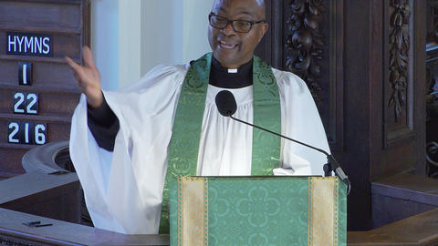 Smiling pastor, the Rev. Emmanuel Akyeampong, in white robe and green stole delivering sermon at church pulpit with hymn numbers displayed on board.