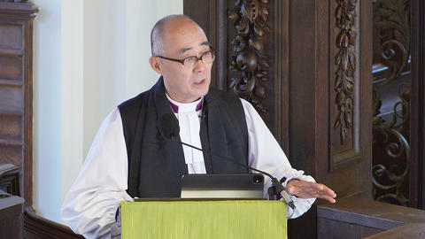 The Rt. Rev. Allen K. Shin, Bishop Suffragan, The Episcopal Diocese of New York, in white robe and black stole speaking at a wooden pulpit with a microphone and green cloth covering.