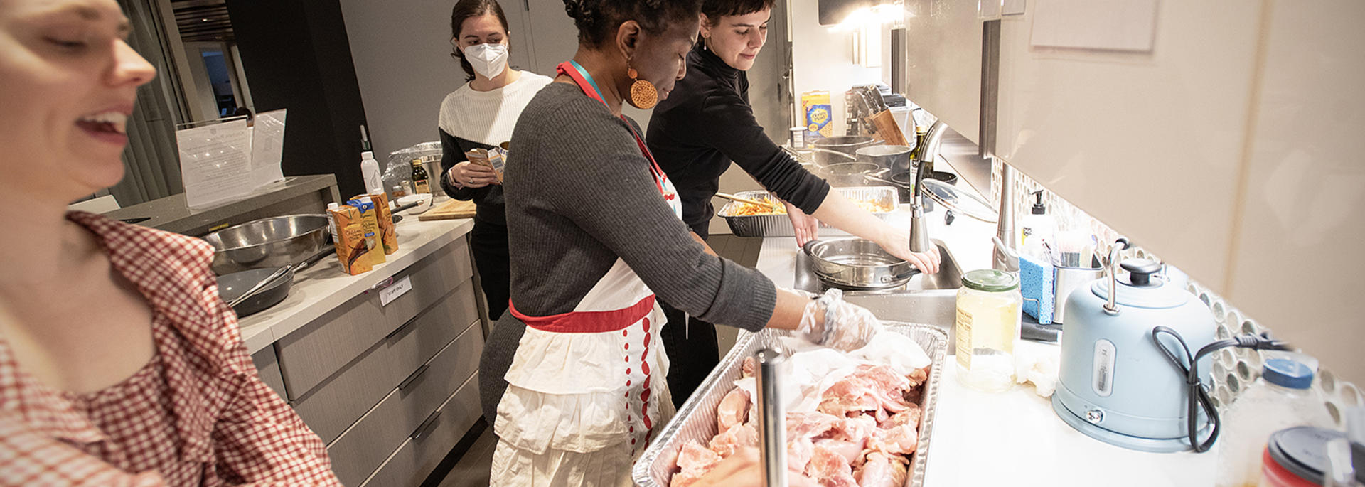 Three women, Anna Burnham, Theresa Lungu, and Elizabeth Propst, preparing raw chicken in a modern kitchen, wearing aprons and gloves for hygiene.